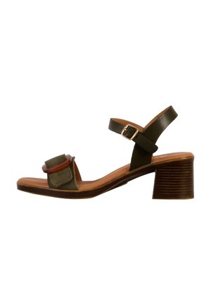 Sandalo con tacco aperto in punta, caratterizzato da una tomaia in suede verde, cinturino in pelle marrone e un tacco in legno spesso con finitura texture. Accentuato da un dettaglio circolare.