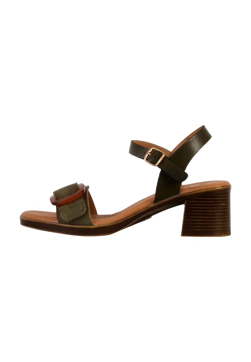 Open-toe sandaal met hak, voorzien van een groene suède bovenkant, een bruine leren band en een chunky houten hak met een gestructureerde afwerking. Accenten met een cirkelvormig detail.
