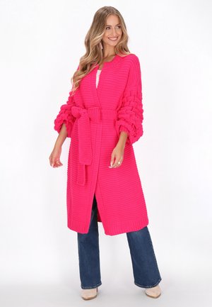 Long pink knitted cardigan with a belt, featuring textured balloon sleeves and an open front design, paired with flared jeans.