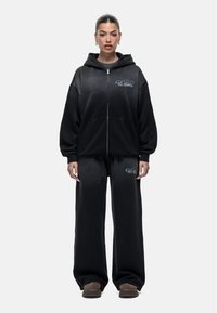 Zwarte capuchontrui en bijpassende joggingbroek, met een ritssluiting aan de voorkant, grafische tekstaccenten en een relaxed fit met wijde benen.
