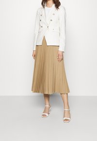 Donna che indossa un blazer bianco doppiopetto, una gonna midi plissettata beige e sandali con tacco aperti bianchi, in piedi contro uno sfondo neutro.