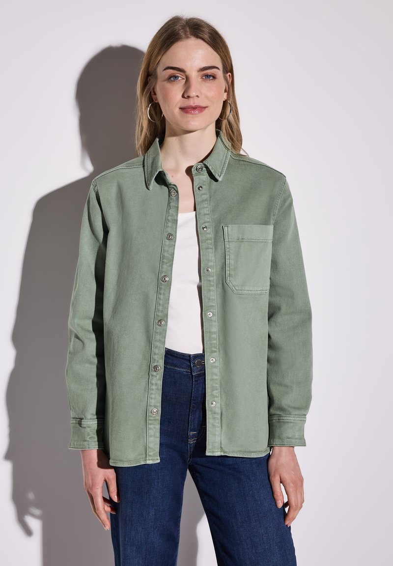 Vaaleanvihreä napillinen paita pehmeästä kankaasta, jossa on rintatasku, yhdistetty valkoiseen yläosaan ja tummansinisiin farkkuihin. Sileä pinta ja rento istuvuus.