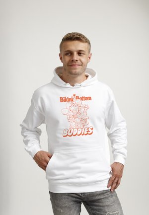 Młody mężczyzna w białej bluzie z kapturem z pomarańczowym napisem „Bikini Bottom Buddies” i postaciami z kreskówek, stojący na tle prostego jasnego tła.
