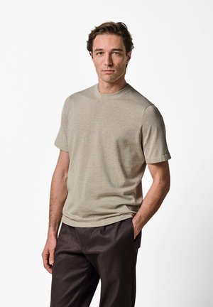 Hombre con la mano en el bolsillo, vistiendo una camisa beige de manga corta y pantalones marrón oscuro, de pie contra un fondo blanco liso.