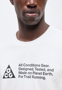 Κοντινό πλάνο ενός ατόμου που φοράει λευκό πουκάμισο με μαύρο κείμενο: «All Conditions Gear. Σχεδιασμένο, δοκιμασμένο και κατασκευασμένο στον πλανήτη Γη. Για τρέξιμο σε μονοπάτια.»