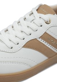 Sneaker sintetico bianco con accenti beige, pannelli laterali texturizzati, lacci bianchi piatti e suola in gomma. Presenta il marchio sul lato.