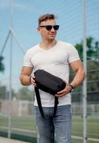 Černá látková crossbody taška s hladkým povrchem a nastavitelným popruhem, kterou drží osoba v bílém tričku a modrých džínách na sportovním hřišti.