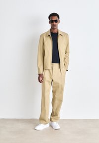 Chaqueta y pantalones beige de corte relajado, combinados con una camisa azul marino y zapatillas blancas. El modelo lleva gafas de sol oscuras.