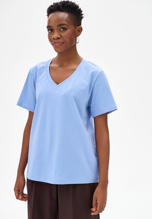 Kobieta o krótkich kręconych włosach, ubrana w jasnoniebieską koszulkę z dekoltem w serek oraz ciemne spodnie, stojąca na jednolitym białym tle.