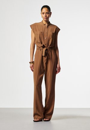 Vrouw staat en draagt een bruine mouwloze jumpsuit met riem, wijde broekspijpen, knopen aan de voorkant, twee borstzakken, armbanden en sandalen op een effen achtergrond.