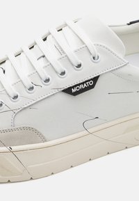 Fehér sneaker fekete és szürke absztrakt mintával. Fehér fűzőkkel, textil felsőrésszel és struktúrált bézs velúr díszítéssel az orrán.