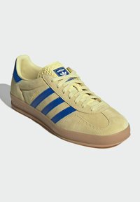 Beige semišové tenisky s modrým třemi pruhy, kulatou špičkou, strukturovanou gumovou podrážkou a pleteným jazykem. Na jazyku logo na modrém pozadí.