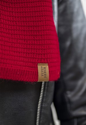 Cappello rosso lavorato a maglia con texture a coste e un'etichetta in pelle beige con "Vytal Sense" sul bordo, indossato con una giacca di pelle nera.