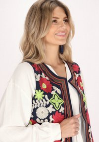 Blommönstrad broderad väst i marinblått med röda, gröna och vita mönster; ärmlös design över en vit blus med rund halsringning.