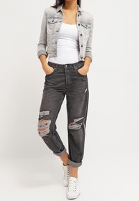 Person med en grå croppad denimjacka, vit linne, svarta trasiga jeans uppvikta vid vristerna och vita sneakers, stående med händerna i fickorna.