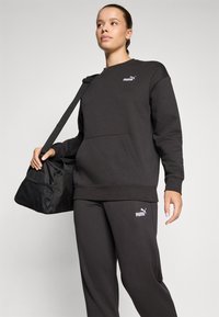 Svart sweatshirt med en framficka och vitt logotyp, i kombination med matchande sweatpants. Personen håller en svart gymväska med nät detaljer.