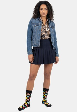 Frau mit lockigem Haar, die eine Jeansjacke, eine gemusterte Bluse, einen marineblauen Faltenrock und schwarze Kniestrümpfe mit bunten Blumenmuster trägt.