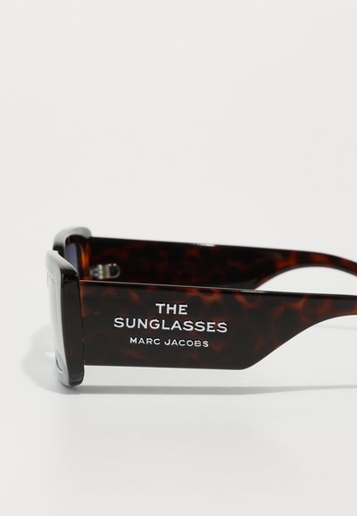 Gafas de sol de carey con montura gruesa. El texto en el costado dice "LAS GAFAS DE SOL MARC JACOBS" en blanco, contrastando con el material oscuro.