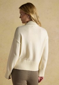 Cremfarbener Pullover mit hochgezogenem Rippkragen, abgesenkten Schultern und geripptem Saum. Weiche Textur, lässige Passform und minimalistisches Design.