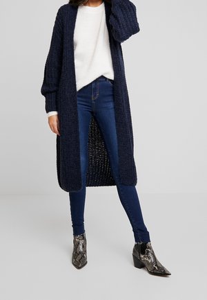 Kvinde iført en lang mørkeblå strikket cardigan, hvid sweater, mørkeblå skinny jeans og ankelstøvler med slangeskindsprint.