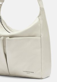 Borsa a spalla in pelle crema con forma curvata, chiusura con zip e due tasche frontali. Presenta un branding minimale nella parte inferiore.