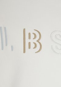 La tela blanca presenta letras bordadas en azul, dorado y blanco, mostrando un diseño moderno y minimalista con formas y texturas variadas.