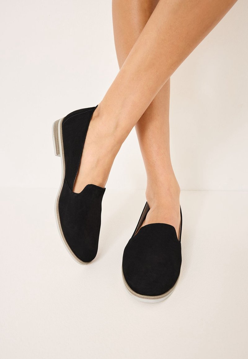 Ballerines noires à enfiler avec semelles claires, portées sur des jambes croisées sur un fond uni clair.