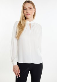 Weiße, langärmlige Bluse mit hohem Kragen und Schlüsselaugen-Detail. Hergestellt aus leichtem Stoff mit glatter Textur. Kombiniert mit schwarzen Hosen.