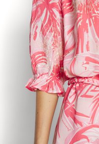 Blouse fleurie rose avec manches à volants, arborant un motif de feuilles de palmier et des paillettes discrètes. Le tissu semble léger et aérien.