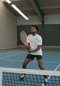 Tennisemängija valges adidas'e särgis ja mustades lühikestes pükstes, hoides oranži ja valget reketit, valmis sinisel väljakul.