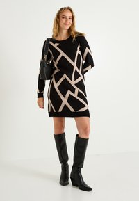 Robe tricotée noire et beige avec un motif géométrique, manches longues, col rond, associée à des bottes noires à talons hauts et un petit sac.
