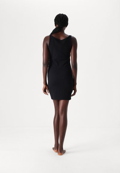 Rochie mini neagră, fără mâneci, cu formă ajustată, textură netedă, decolteu rotund și detalii pe spate. Purtați de o persoană desculță.
