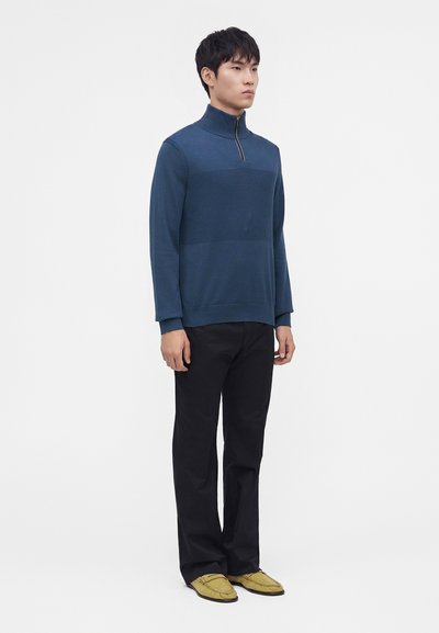 Homme debout portant un pull bleu à fermeture éclair au quart, un pantalon noir et des chaussures jaunes sans lacets sur un fond clair uni.