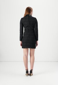 Ein Model trägt ein enges schwarzes Kleid mit strukturiertem Muster, zeigt den Rücken und kombiniert es mit schwarzen High Heels vor einem weißen Hintergrund.
