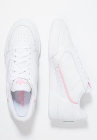 Sneaker bianche con una texture in pelle liscia, caratterizzate da accenti rosa, dettagli forati sulla punta e un'etichetta con il marchio sulla linguetta.