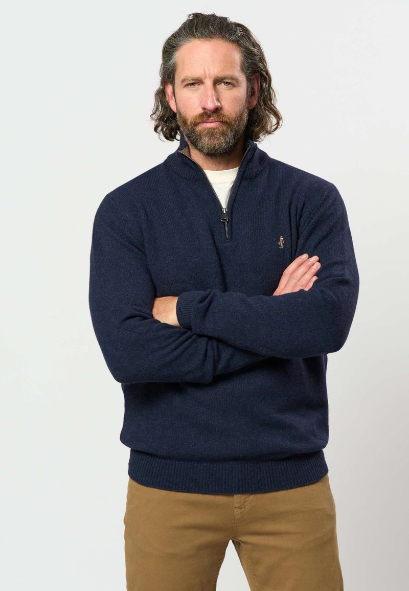 Hombre con barba y cabello ondulado, de pie con los brazos cruzados, vistiendo un suéter azul marino con media cremallera y pantalones marrones sobre un fondo liso.