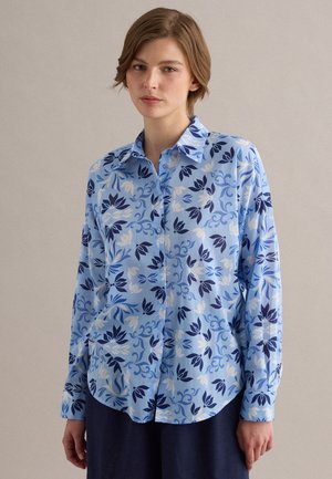 Donna con i capelli corti che indossa una camicia azzurra con bottoni e motivo floreale blu scuro e bianco, in piedi contro uno sfondo chiaro e semplice.