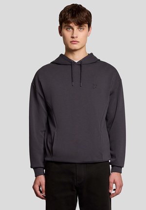 Jonge man met bruin haar, gekleed in een donkergrijs capuchontrui en zwarte broek, staand tegen een effen achtergrond.