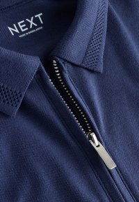 Col polo à fermeture éclair bleu marine avec fermeture éclair argentée, tissu texturé et étiquette "NEXT Fabriqué au Bangladesh" à l'intérieur du col.