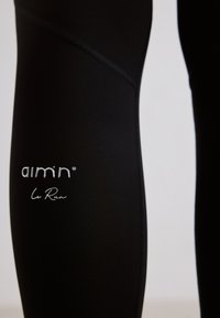 Gros plan sur un legging de sport noir avec des coutures surpiquées et le logo "aim'n le Run" imprimé sur la cuisse.