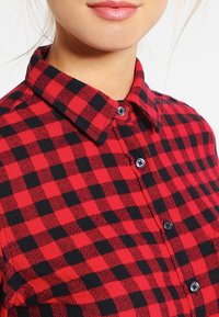 Röd och svart rutigt flanellskjorta med knappat krage och svarta knappar, med en mjuk texturerad tyg.