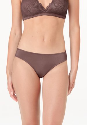 Donna che indossa un bralette in pizzo marrone e slip bikini marroni abbinati, in piedi contro uno sfondo bianco uniforme.