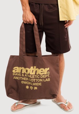 Mann hält große braune Tragetasche mit gelbem Text, trägt dunkelbraune Shorts, gelbes Shirt und weiße Flip-Flops vor hellem Hintergrund.