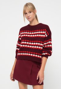 Knit pullover sweater in burgundy with red and white heart patterns, featuring a round neck and slouchy sleeves, paired with a burgundy skirt.