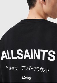 Personne portant un t-shirt noir avec un grand texte blanc "ALLSAINTS" et des caractères japonais plus petits au-dessus de "LONDON" dans le dos.
