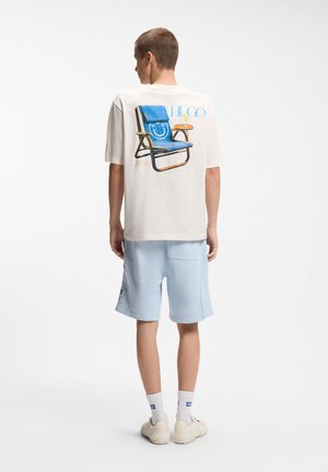 Man wearing white T-shirt with blue chair graphic and light blue shorts, standing facing away against white background.