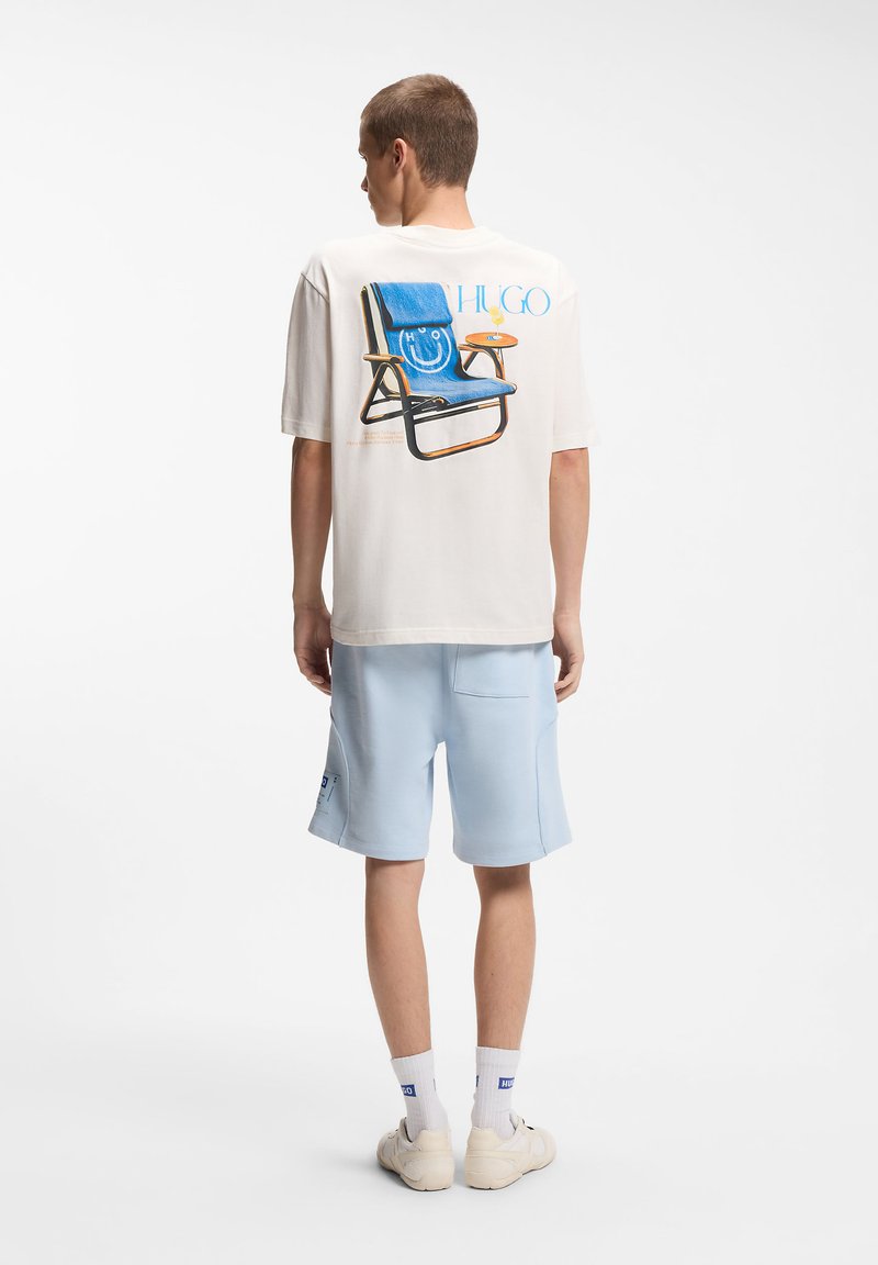 Άνδρας που φοράει λευκό μπλουζάκι με μπλε γραφικό καρέκλας και ανοιχτό μπλε σορτς, στέκεται με την πλάτη προς τα εμπρός σε λευκό φόντο.