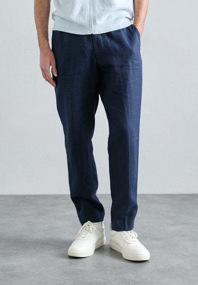 Pantalones de lino mezclado en color navy con un corte relajado y pliegues visibles. Combinados con zapatillas blancas, sobre un fondo gris claro.