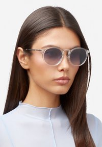 Okrugle sunčane naočale s prozirnim okvirom, gradijentnim lećama i metalnim detaljima. Model nosi svijetloplavu majicu s visokim ovratnikom.