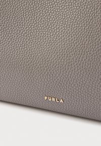 Šedá kožená taška s texturovaným povrchem a zlatým logem "Furla". Má obdélníkový tvar a nabízí sofistikovaný vzhled.
